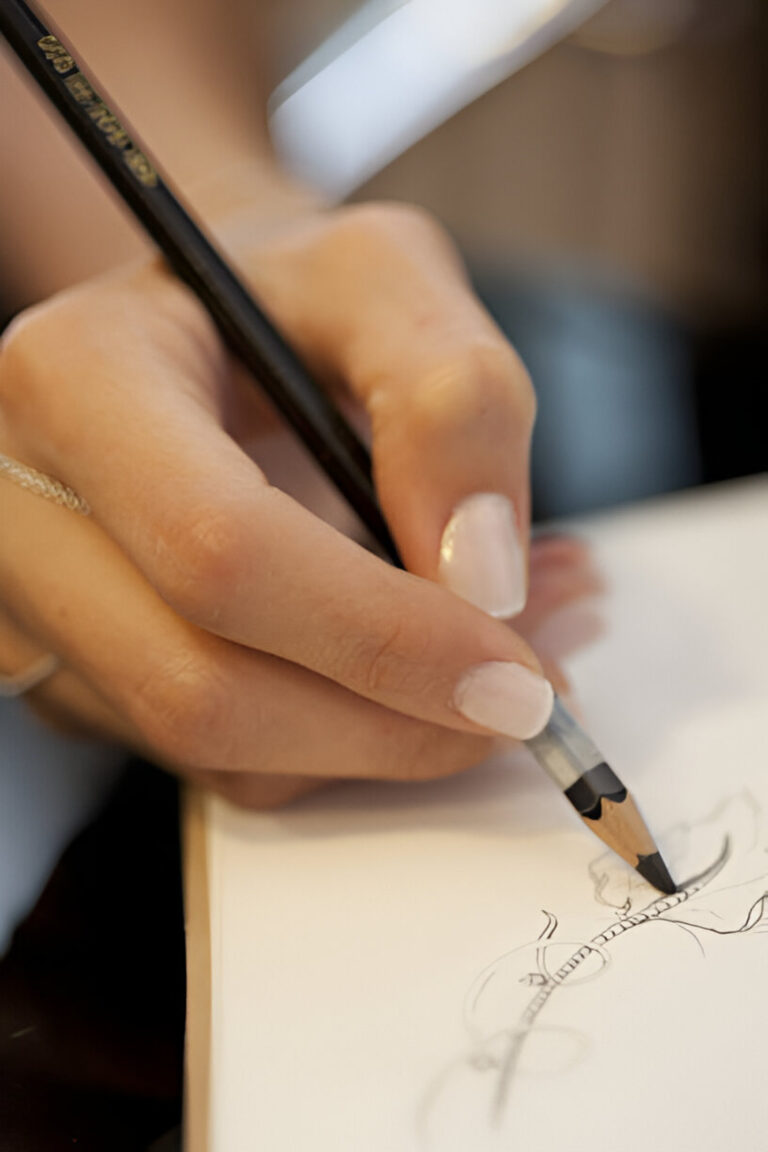 Close-up of jewelry designer's manicured hands holding pencil while sketching multiple jewelry design iterations on white paper, showing detailed ring concepts and variations