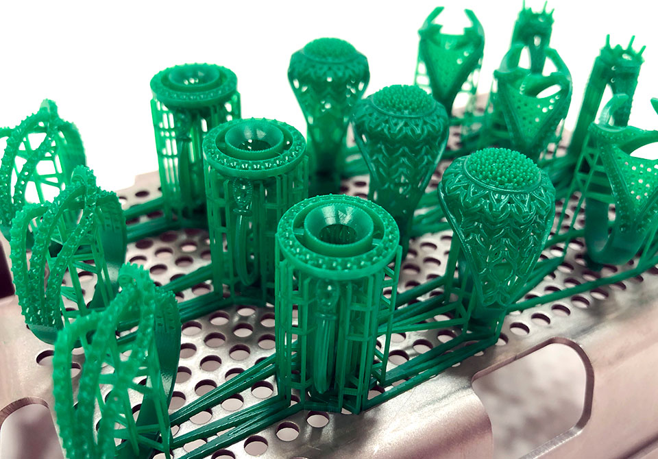 Green 3D printed castable wax jewelry models on 3D printer build platform with perforated surface ready for casting